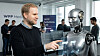 En mann peker på en humanoid robot foran en skjerm i et lyst kontor.