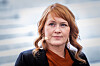 Portrett av en kvinne med rødt hår, mørk jakke og oransje bluse foran en lys bakgrunn.