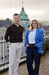 To personer står på en takterrasse foran byens hustak og en grønnkupplet bygning.