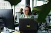 Inger-Marit ved laptop på kontoret med skjermer, planter og vinduer.