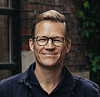 Smilende person med briller foran en murvegg og grønne planter.
